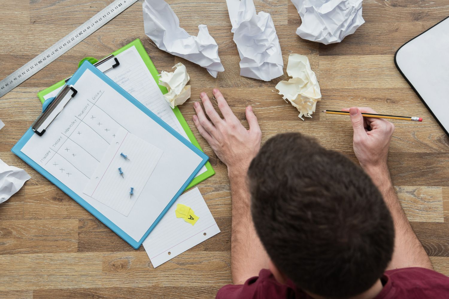 Homem visto de cima, cercado por folhas de papel amassadas e planilhas sobre uma mesa, indicando tentativas frustradas de organização. A imagem remete aos desafios da gestão da equipe de recepção, destacando a necessidade de mapear erros recorrentes para tornar os processos mais eficientes e funcionais.