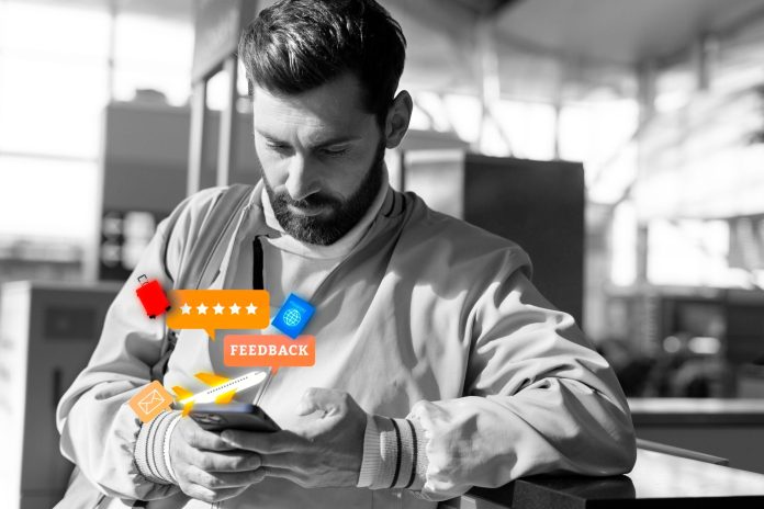 Homem com expressão concentrada mexe no celular em um ambiente que remete a uma recepção de hotel, com ícones digitais coloridos de avaliação, feedback e viagem sobrepostos à imagem. A cena ilustra a importância de monitorar o desempenho da equipe da recepção, que lida diretamente com a experiência do hóspede.