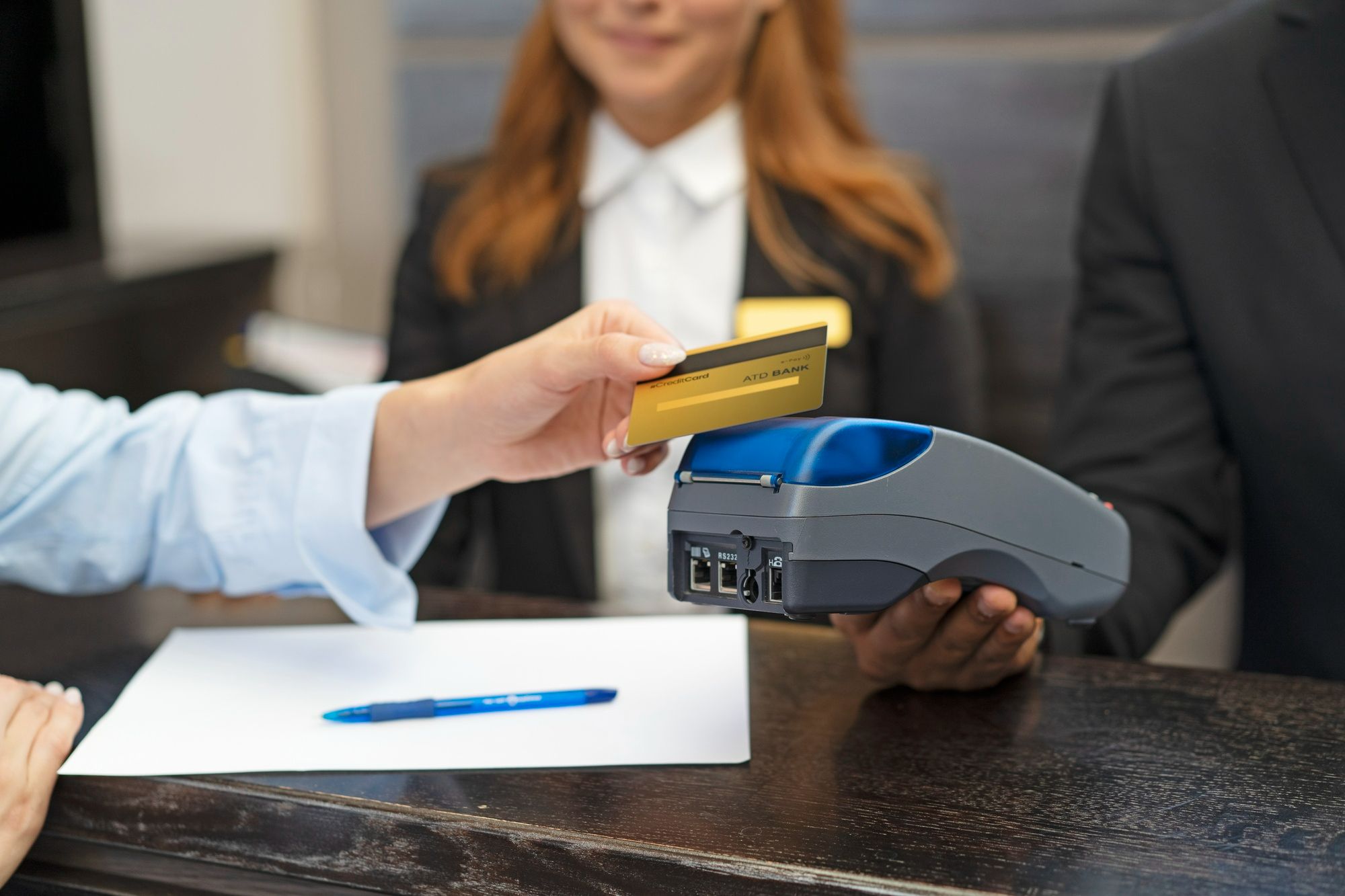 Hóspede realiza pagamento com cartão na recepção de um hotel, enquanto colaborador segura a máquina de débito/crédito. A imagem representa o papel estratégico da recepção na geração de receita, com destaque para práticas de upselling e cross-selling.