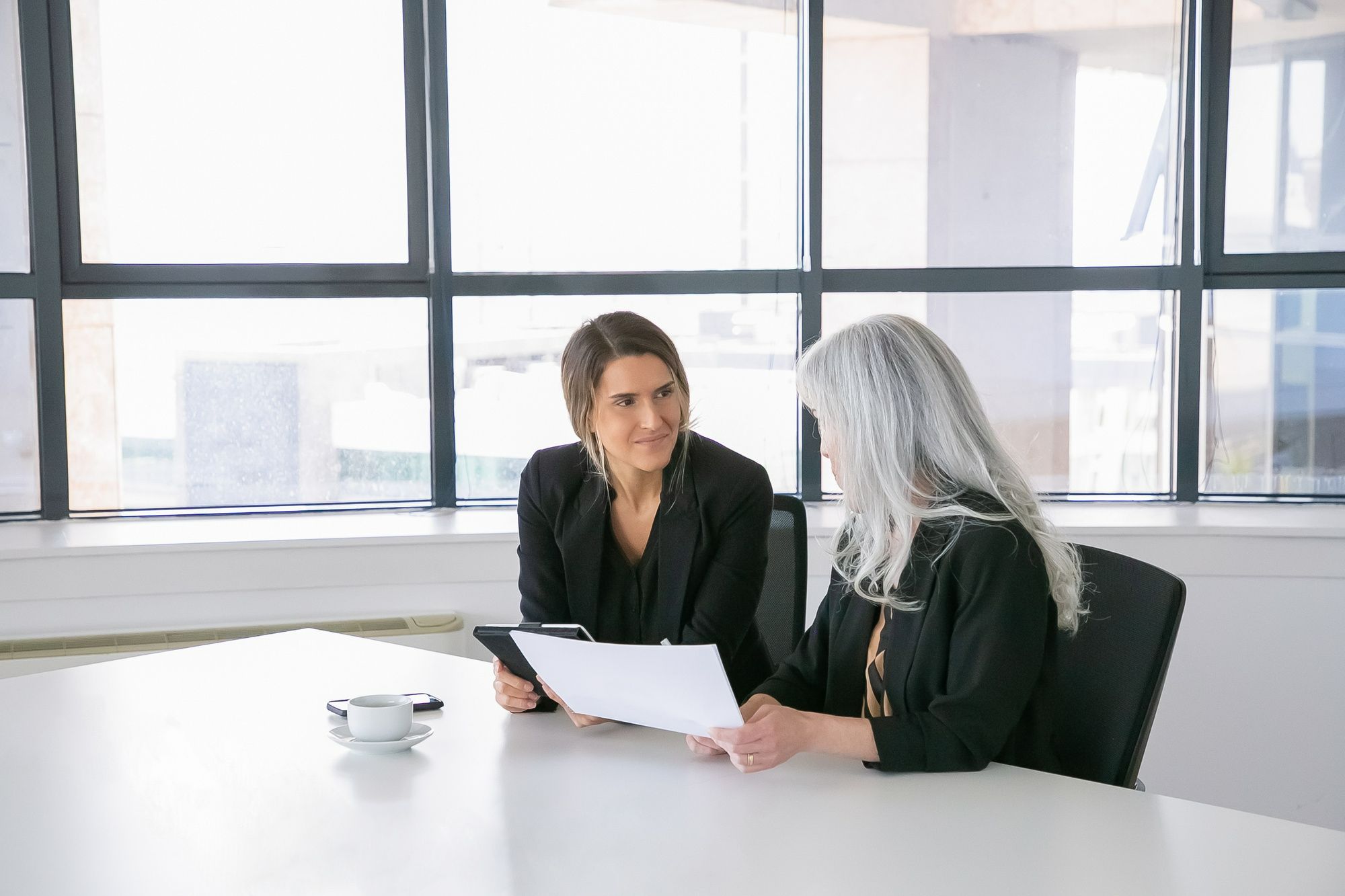 Duas profissionais sentadas à mesa durante uma conversa. Uma segura um tablet e observa a colega, que analisa um documento impresso. A imagem remete à importância do feedback construtivo e da confiança mútua na gestão do desempenho da equipe da recepção.