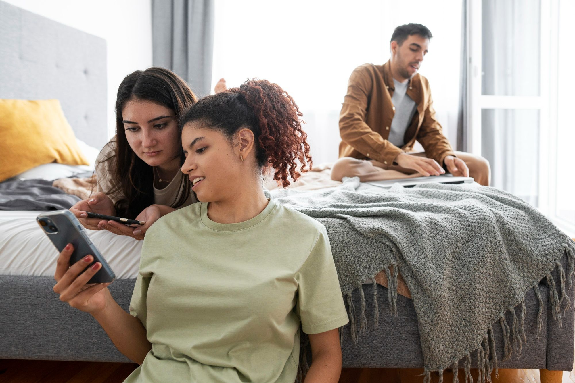 Três pessoas em ambiente descontraído, duas delas olhando juntas o celular enquanto a terceira usa um notebook ao fundo. A imagem ilustra como definir o público-alvo é um passo essencial para impactar as pessoas certas (no caso, por meio do smartphone e do computador), contribuindo para o sucesso dos pacotes de hospedagem para a baixa temporada.