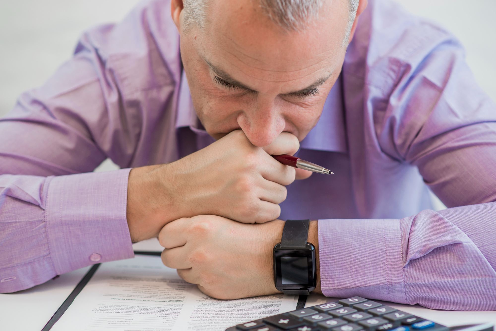 Homem de camisa social roxa, com expressão preocupada, analisa documentos ao lado de uma calculadora. A imagem ilustra a importância de estudar o mercado com atenção antes de negociar e reforça uma das principais dicas de negociação para hotéis.