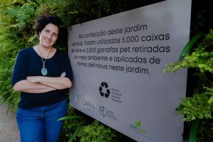 A arquiteta paisagista Maria Paula Orlando, de cabelos curtos e cacheados, usa blusa preta e jeans e posa sorrindo com os braços cruzados ao lado de uma placa informativa instalada em um jardim vertical. A placa explica que o espaço foi construído com 5 mil caixas de leite e 3 mil garrafas PET reaproveitadas, como parte de um projeto sustentável de paisagismo na hotelaria desenvolvido para o Cyan Resort by Atlantica, em Itupeva (SP).