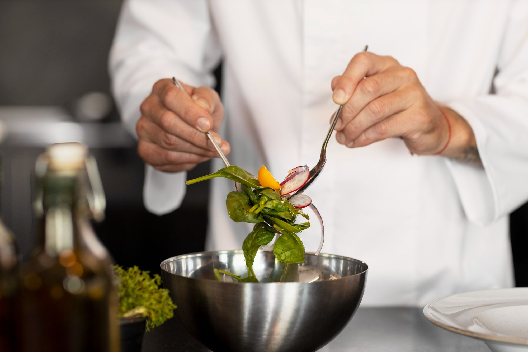 Chef de cozinha, vestindo jaleco branco, prepara cuidadosamente uma salada fresca em uma tigela de inox. A composição inclui folhas verdes, rabanetes fatiados e um pedaço de tomate. A cena remete à valorização de práticas sustentáveis e à oferta de uma alimentação saudável na hotelaria.