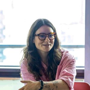 Andreza Ranieri, gerente de Responsabilidade Social Corporativa da Accor para as Américas, aparece sentada em uma cadeira vermelha, com expressão serena e um leve sorriso. Ela usa óculos de armação tartaruga, camisa rosa de mangas dobradas e um relógio preto no pulso esquerdo, que também exibe uma tatuagem delicada de flores.