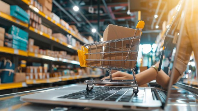 Carrinho de compras em miniatura com caixa de papelão dentro, posicionado sobre um teclado de notebook, em frente a prateleiras de almoxarifado com materiais organizados. A imagem simboliza o gerenciamento estratégico de estoque para eventos em hotéis, destacando a importância da digitalização e do controle eficiente de insumos para otimizar custos e fortalecer a imagem do meio de hospedagem.