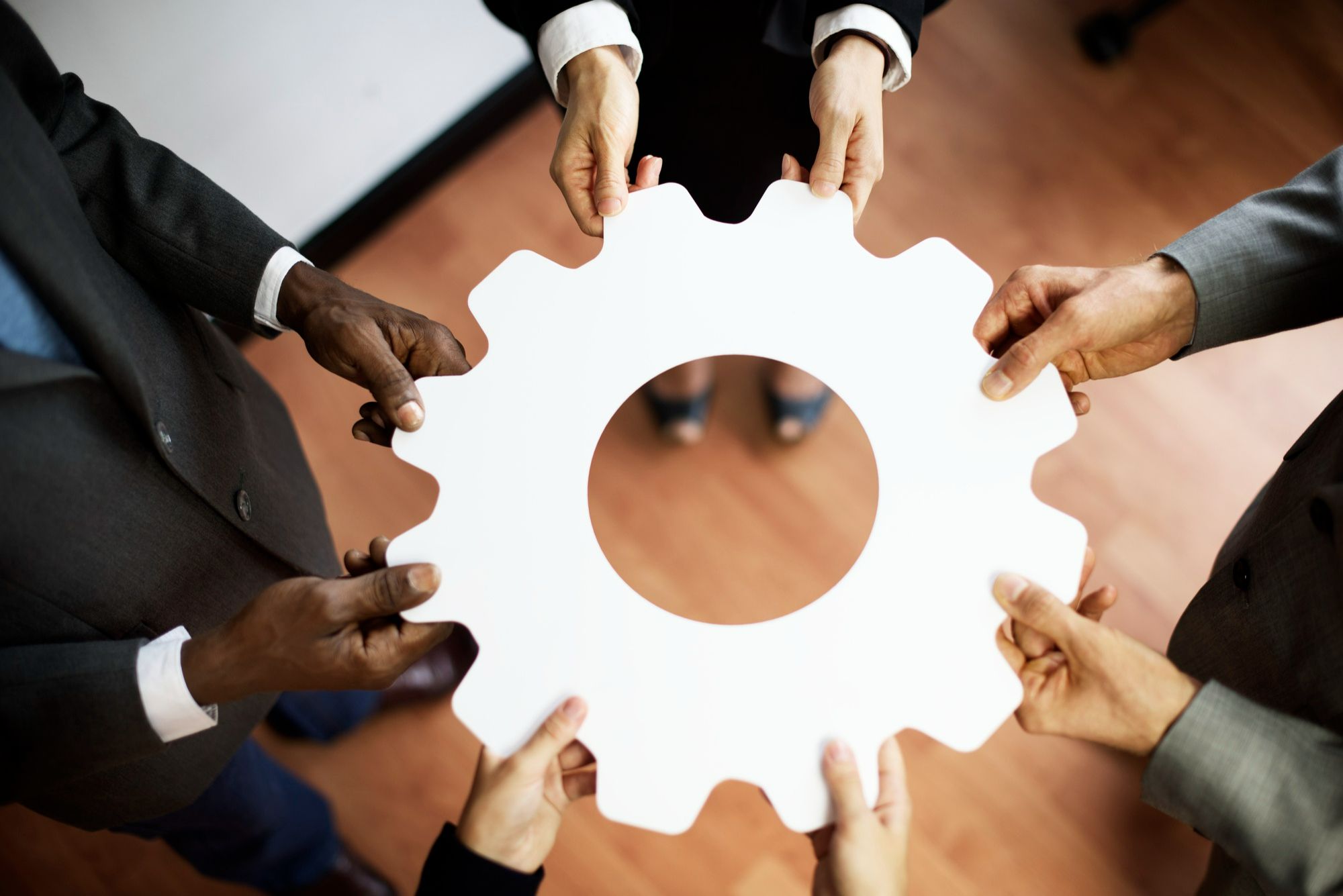 Pessoas de diferentes perfis seguram, em conjunto, uma engrenagem branca, simbolizando como a gestão de crises na hotelaria se fortalece com a atuação colaborativa e multidisciplinar das equipes.