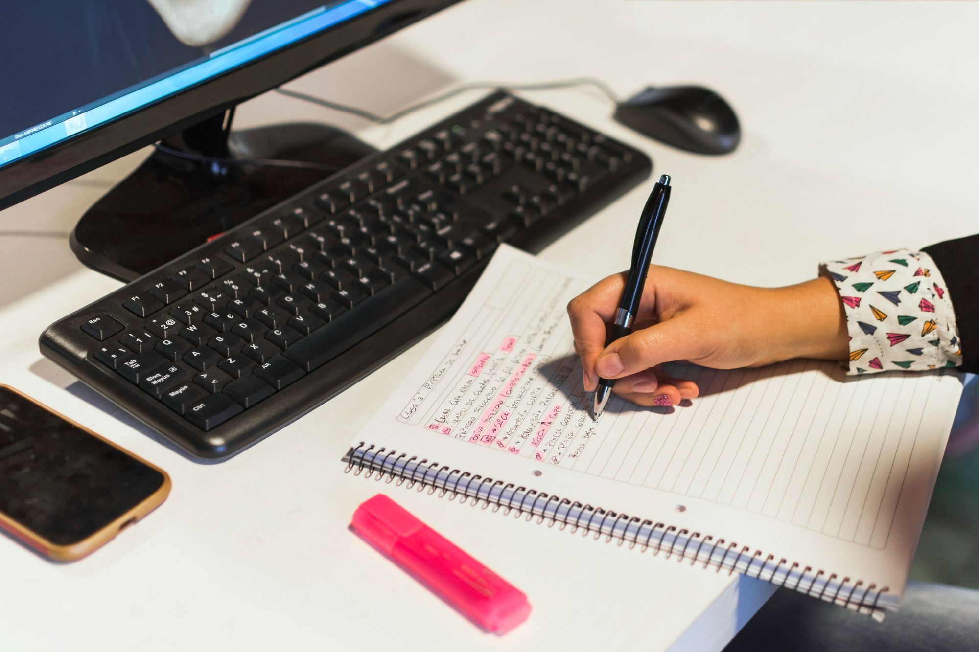 Pessoa escrevendo anotações em um caderno ao lado de computador, celular e marca-texto, representando o uso de scripts comerciais como ferramenta de apoio para garantir um atendimento de alto padrão na hotelaria.