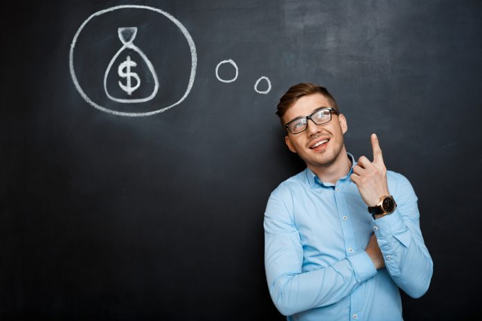 Homem sorridente, em frente a um quadro-negro, apontando para cima, em direção a um desenho de um saco de dinheiro, simbolizando ideias lucrativas. A imagem ilustra como o atendimento de alto padrão na hotelaria pode gerar mais receita para o hotel.