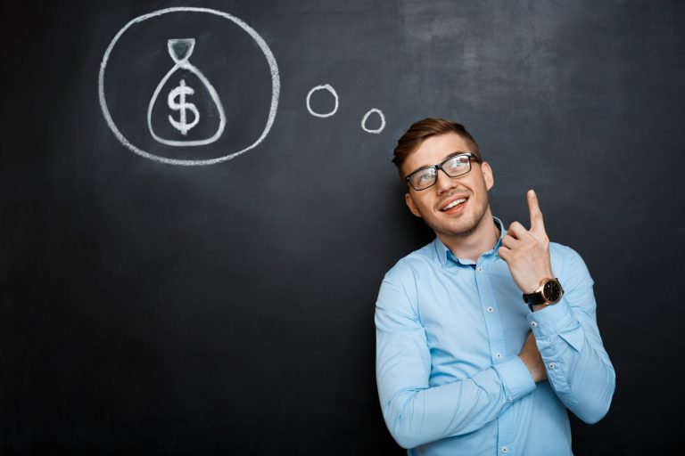 Homem sorridente, em frente a um quadro-negro, apontando para cima, em direção a um desenho de um saco de dinheiro, simbolizando ideias lucrativas. A imagem ilustra como o atendimento de alto padrão na hotelaria pode gerar mais receita para o hotel.