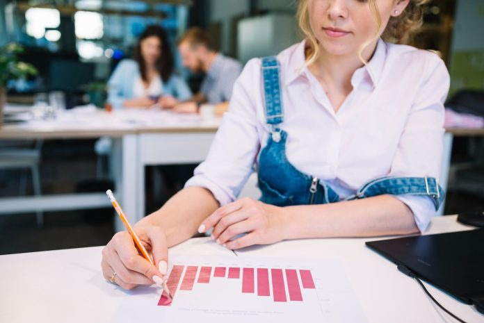 Mulher analisa gráfico de barras impresso, usando lápis para destacar informações, enquanto trabalha em um ambiente corporativo com colegas ao fundo. A cena representa a importância da análise de métricas para avaliar o sucesso de eventos em hotéis, indo além da contagem de participantes.
