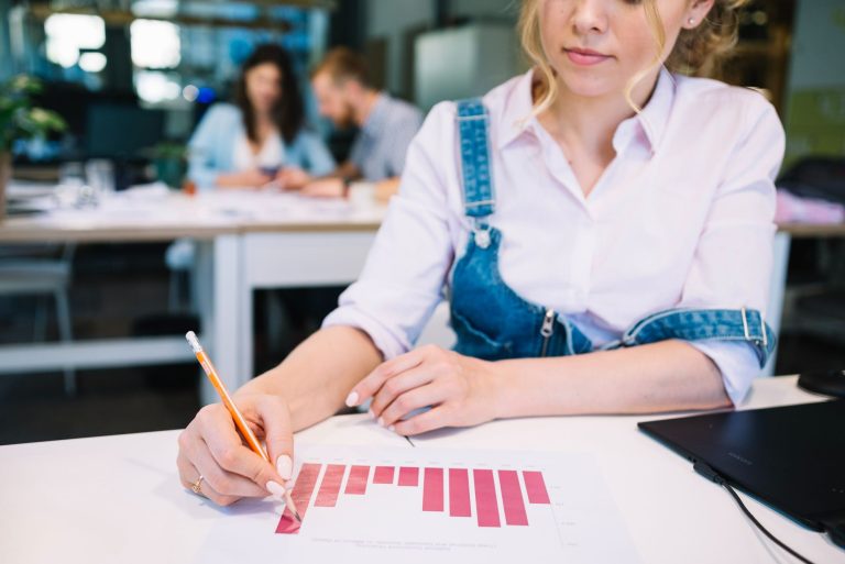 Mulher analisa gráfico de barras impresso, usando lápis para destacar informações, enquanto trabalha em um ambiente corporativo com colegas ao fundo. A cena representa a importância da análise de métricas para avaliar o sucesso de eventos em hotéis, indo além da contagem de participantes.