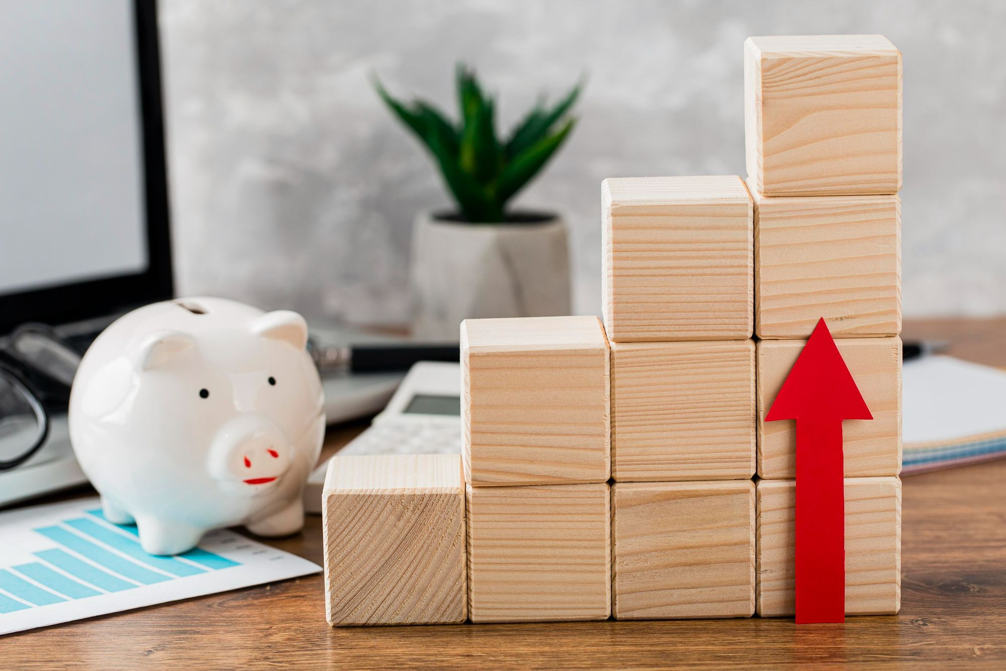 Cofrinho branco ao lado de gráfico financeiro azul, com blocos de madeira formando uma escada crescente e uma seta vermelha apontando para cima. A imagem simboliza o balanço financeiro como uma das métricas essenciais para avaliar o sucesso de eventos em hotéis, destacando a importância do crescimento e da rentabilidade.
