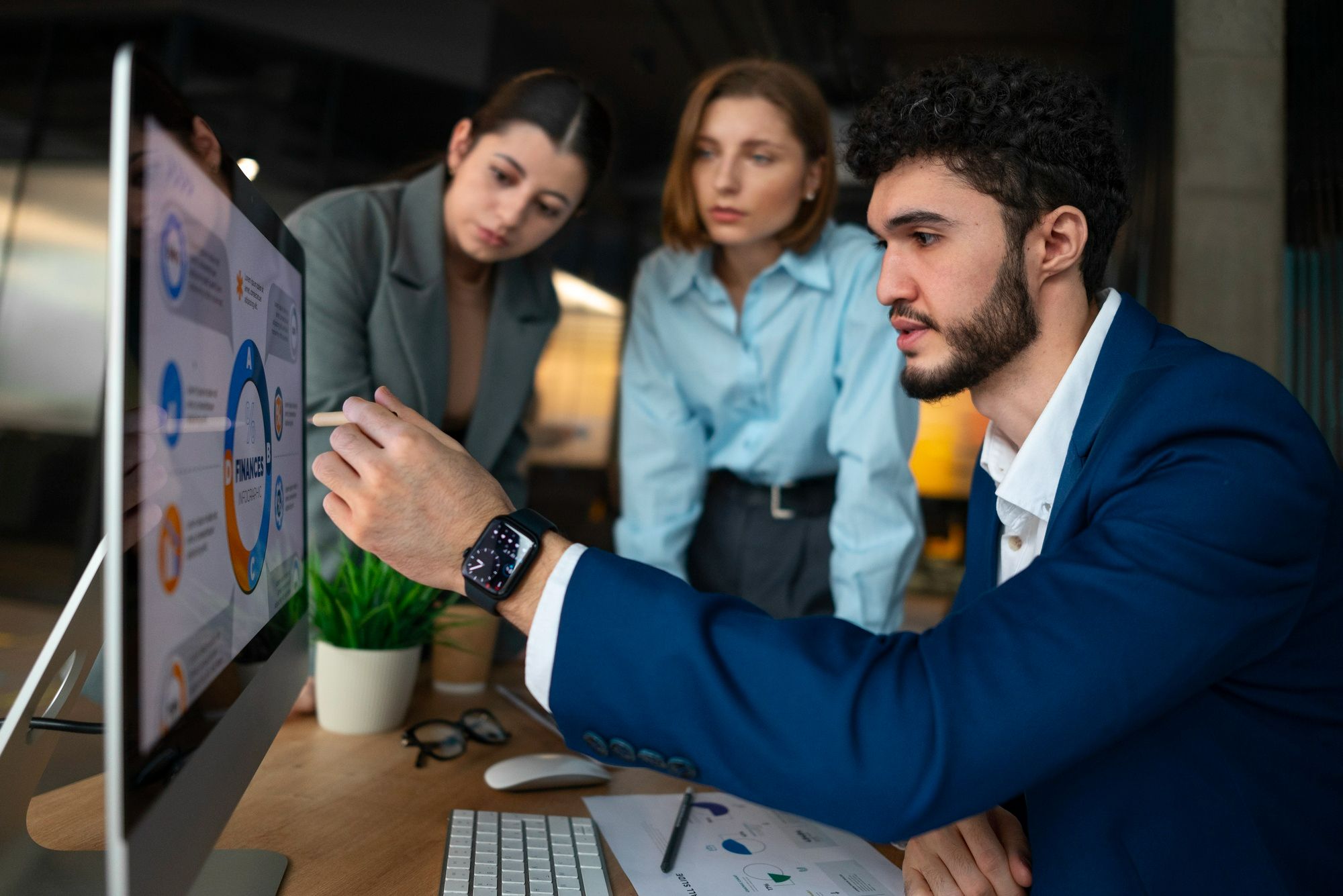Profissional aponta para gráficos em uma tela de computador enquanto apresenta dados a duas colegas durante uma reunião; a cena destaca a importância do raciocínio analítico e da interpretação correta das informações para uma gestão de compras com big data eficiente.