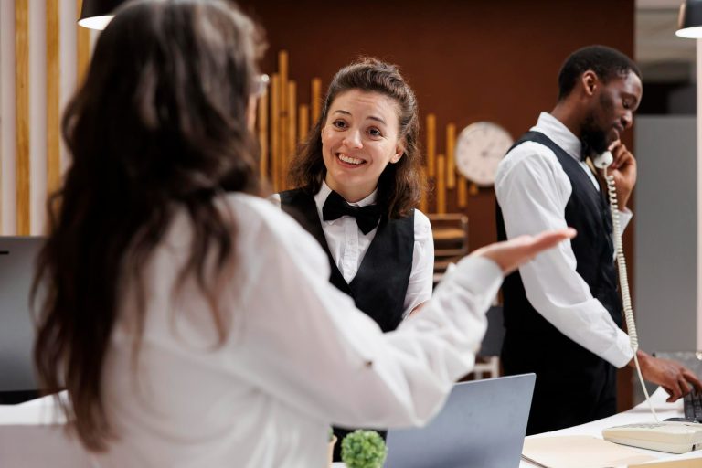 Recepcionistas de hotel interagem de forma cordial e atenciosa na área da recepção; uma profissional sorri enquanto conversa com uma colega, enquanto outro funcionário atende ao telefone ao fundo. A imagem ilustra a importância da inteligência emocional na recepção hoteleira para garantir uma experiência positiva ao hóspede.