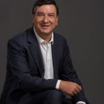 Homem de terno azul-marinho e camisa branca, sentado e sorrindo em frente a um fundo neutro escuro – retrato corporativo de Jeferson Munhoz, CEO da Escola para Resultados.