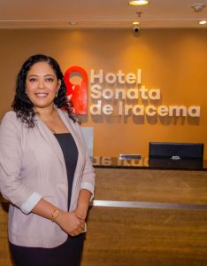 Retrato de Raquel Pinheiro, uma mulher morena e sorridente, posando em pé com as mãos juntas à frente do balcão de recepção de um hotel com iluminação quente e o logotipo "Hotel Sonata de Iracema" ao fundo.