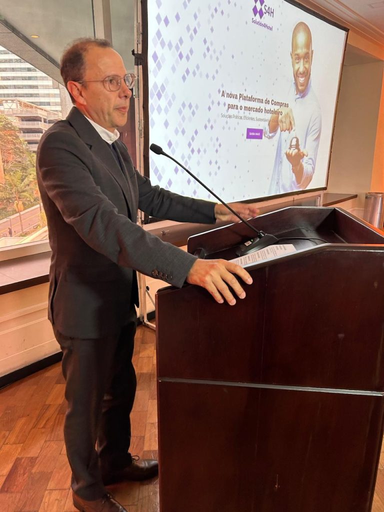 Marcos Vilas Bôas, presidente da ABIH-SP, discursa em púlpito durante evento sobre retrofit tecnológico em hotéis. Ao fundo, projeção da plataforma Solution4Hotel destaca soluções práticas, eficientes e sustentáveis para o mercado hoteleiro, com foco em compras colaborativas.