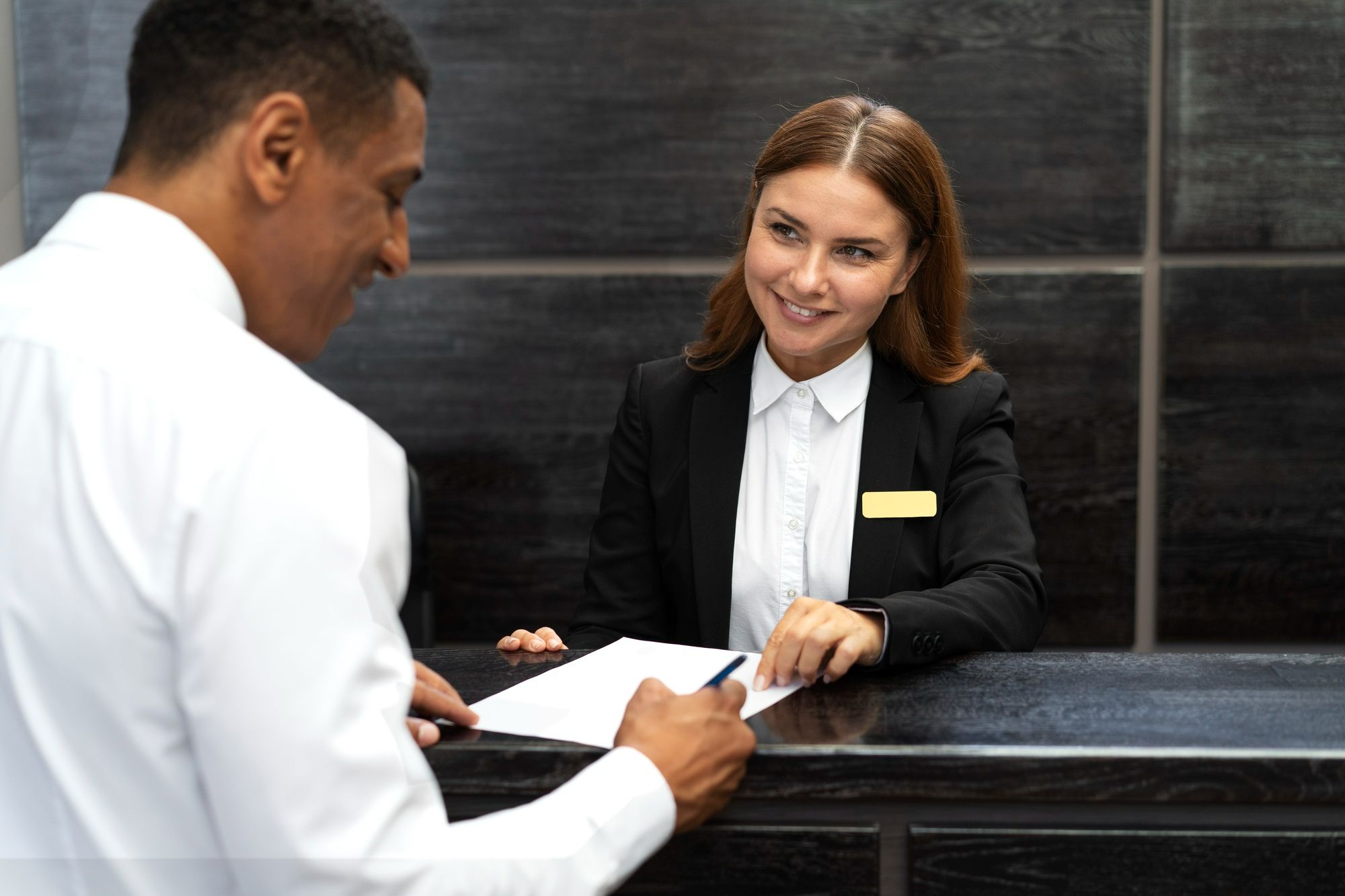 Uma sorridente recepcionista de hotel, vestindo um blazer preto e com um crachá de identificação, está atrás de um balcão, olhando com atenção para um hóspede que assina um documento. A imagem mostra a interação positiva entre o funcionário e o cliente, ilustrando a importância de um bom atendimento e de colocar a experiência do cliente em primeiro lugar para lidar com problemas técnicos em hotéis.