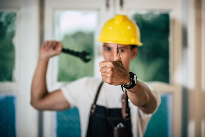 Um técnico de manutenção, usando um capacete de segurança amarelo e um avental de couro, segura uma chave inglesa na mão direita e faz um sinal de 