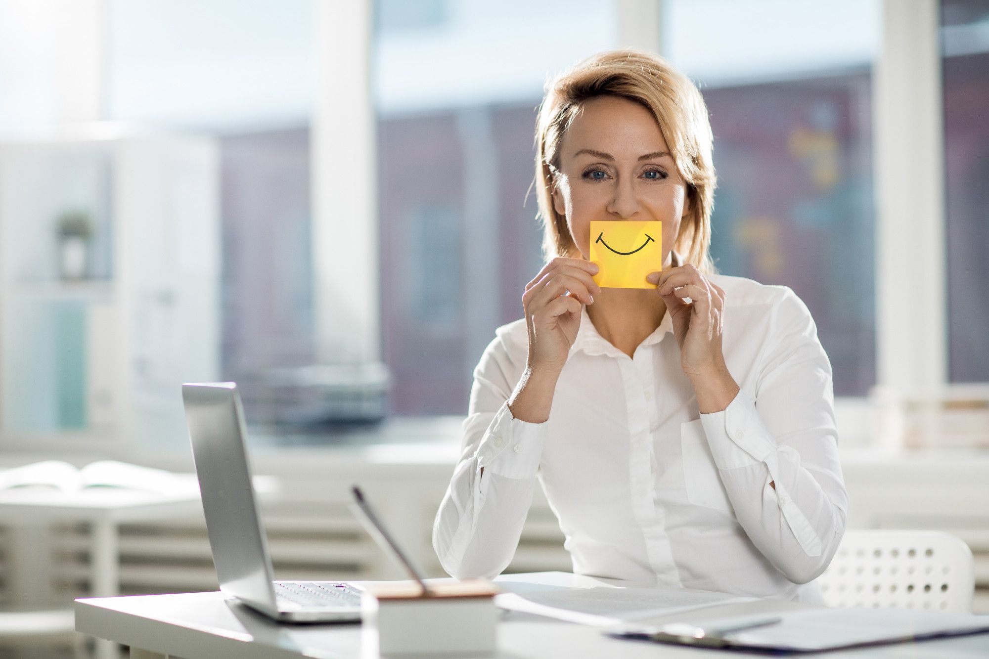 Uma mulher loira de camisa branca, sentada à mesa de um escritório com um laptop, segura um post-it amarelo com um rosto sorridente pintado na frente da boca. A imagem sinaliza que é possível se aperfeiçoar na arte da comunicação não verbal na hotelaria a fim de proporcionar um atendimento de excelência.