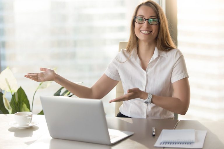 Uma jovem sorridente, usando óculos e mantendo contato visual, está sentada a uma mesa de escritório com um laptop, uma xícara de café e um bloco de notas. Ela tem os ombros relaxados e gesticula com as palmas das mãos abertas, como se apresentasse algo ou indicasse para onde alguém deve ir. A foto ilustra a importância da linguagem corporal na recepção de hotéis e seu papel na criação de uma experiência positiva para o visitante.
