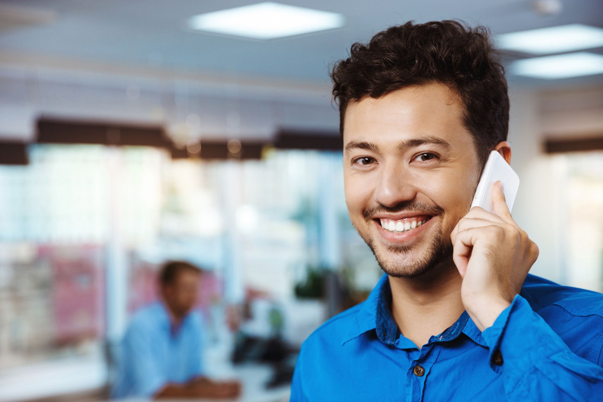 Um homem de cabelo e barba curtos, sorridente e com uma camisa azul, segura um smartphone na orelha. A imagem, com foco no homem e fundo desfocado de um ambiente de escritório, exemplifica a importância do sorriso na comunicação não verbal para criar uma impressão amigável e acolhedora na hotelaria.