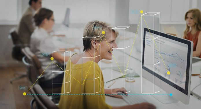 Mulher sorridente de cabelos curtos e grisalhos, vestindo blusa amarela, sentada em frente a um computador desktop em um escritório moderno. No monitor, gráficos e dados financeiros são exibidos. Sobre a imagem, há uma sobreposição gráfica com gráficos de barras tridimensionais e uma linha pontilhada amarela conectando pontos, representando o uso de BI na hotelaria. Ao fundo, outras pessoas trabalham em seus computadores, desfocadas.