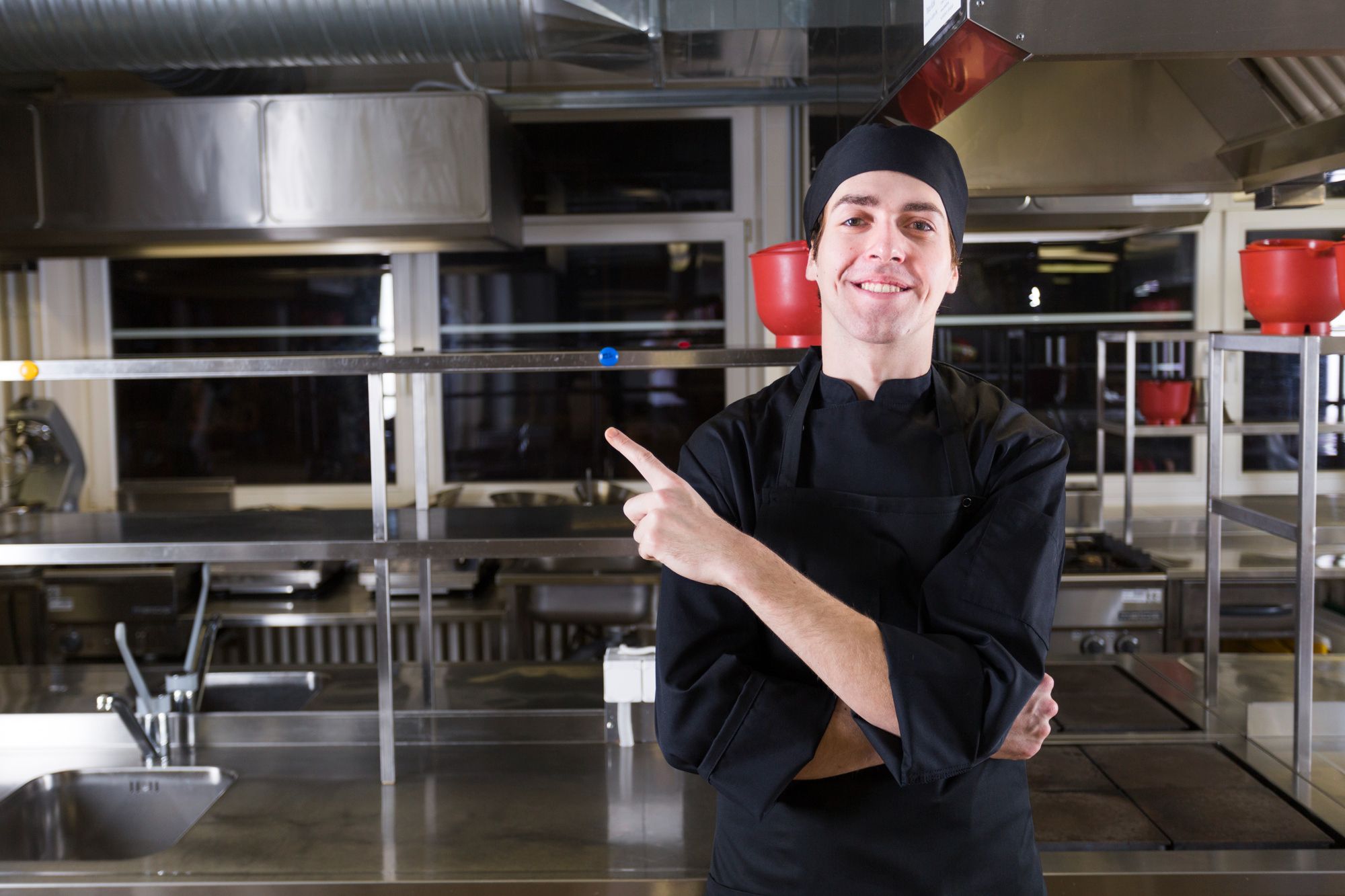 Um jovem cozinheiro sorridente, vestindo um uniforme preto completo com touca preta cobrindo os cabelos, está em pé com os braços cruzados em uma cozinha de aço inoxidável bem iluminada. Ele aponta com o dedo indicador da mão direita para a esquerda do quadro. Ao fundo, observa-se uma bancada com uma pia à esquerda e outros equipamentos de cozinha em inox, além de alguns utensílios vermelhos. A cena ilustra a importância do uso correto de uniforme e dos EPIs para a segurança alimentar em ambientes profissionais.
