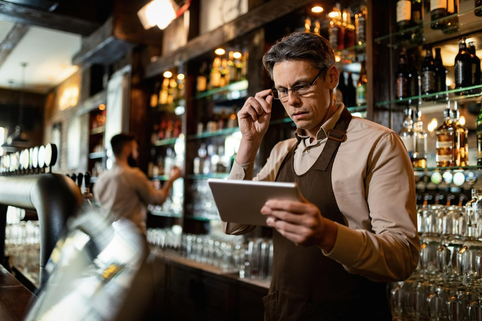 Um homem de óculos, vestindo avental e camisa, está em pé em um bar de restaurante, segurando um tablet e ajustando os óculos com a mão. A imagem, com um colega de trabalho desfocado ao fundo, ilustra a figura de um gestor de A&B inspecionando a operação e servindo de exemplo na rotina que envolve a segurança alimentar em hotéis.