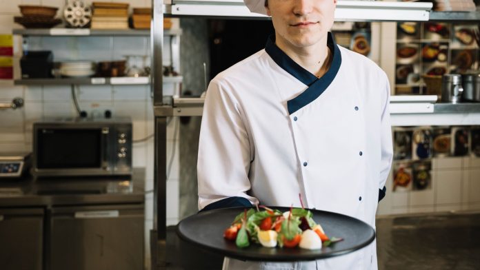 Chef de uniforme branco e chapéu segura prato com salada colorida em uma cozinha profissional, simbolizando boas práticas de segurança alimentar em hotéis.