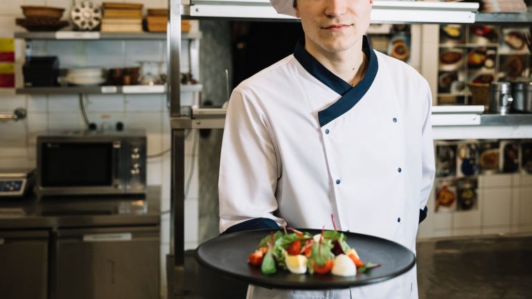 Chef de uniforme branco e chapéu segura prato com salada colorida em uma cozinha profissional, simbolizando boas práticas de segurança alimentar em hotéis.