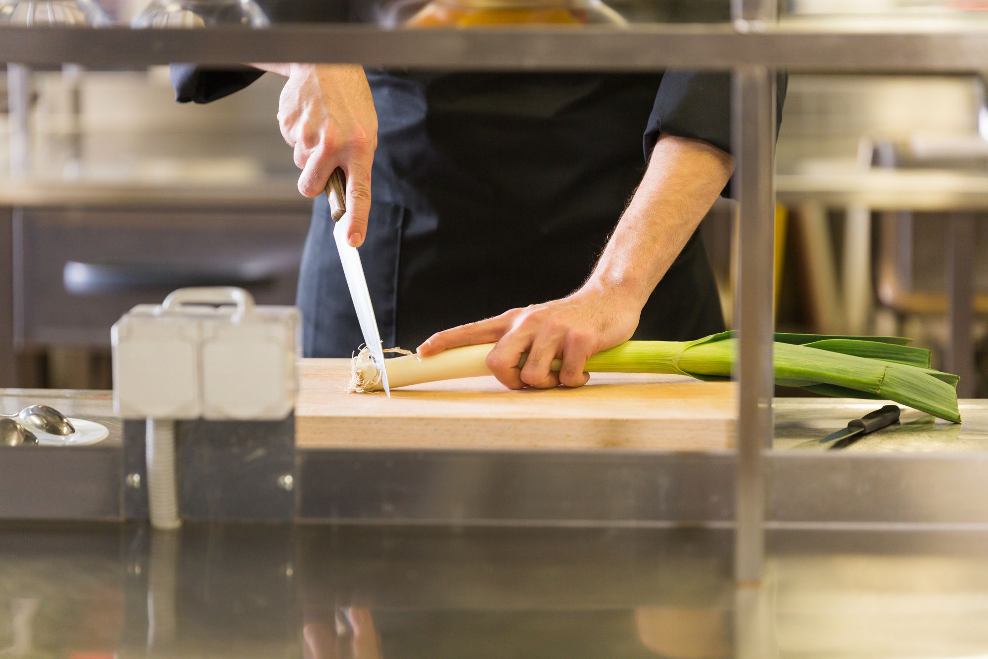 Um close de um cozinheiro em uma cozinha profissional de aço inoxidável, com as mãos segurando um alho-poró em uma tábua de madeira enquanto o corta com uma faca grande. A imagem foca no manuseio do alimento, destacando a importância da higiene e do uso de utensílios na prevenção da contaminação cruzada.