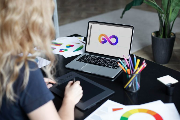 Uma mulher loira sentada em uma mesa de escritório, de costas para a câmera, usa uma mesa digitalizadora com uma caneta. Em seu laptop, na tela, está o que parece ser uma ferramenta de design gráfico exibindo um logotipo de um símbolo do infinito com cores vibrantes. Sobre a mesa, há papéis com rascunhos de designs e um estojo com lápis de cor. A imagem ilustra o processo criativo no desenvolvimento de uma identidade visual, um passo essencial para quem deseja construir uma marca forte na hotelaria.