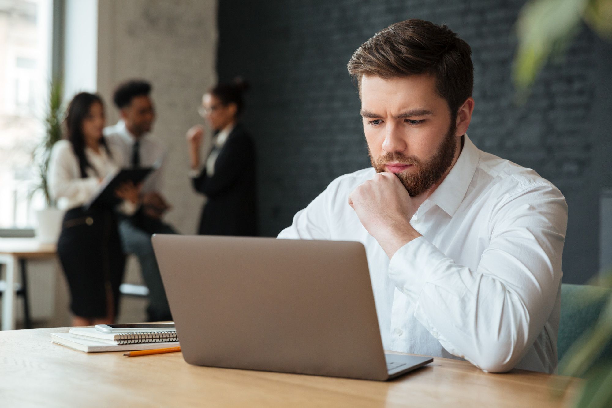 Um homem barbudo com uma camisa branca está sentado pensativamente em uma mesa de madeira, olhando para um laptop aberto. Sua mão direita apoia o queixo. Ao fundo, desfocados, estão três colegas de trabalho interagindo em um ambiente de escritório moderno com paredes de tijolos aparentes. A cena ilustra a importância da concentração e do trabalho individual, que, quando alinhados com sistemas atualizados, contribuem para evitar falhas de comunicação em hotéis através da imediata atualização de informações de reserva.