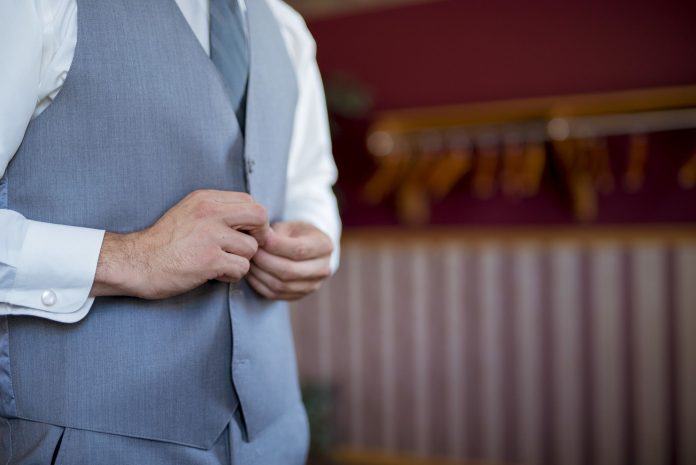 As mãos de um homem que veste um colete e uma camisa branca estão ajustando os botões do colete em um close. Ele também usa uma gravata azul-clara, parcialmente visível acima do colete. O foco está nas mãos e na parte superior do torso, com o fundo ligeiramente desfocado. A cena evoca a preparação e o cuidado com a aparência, elementos importantes na escolha de uniformes para a hotelaria que combinem elegância e praticidade.