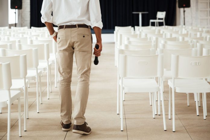 Um homem de costas, vestindo calças cáqui, camisa branca com as mangas dobradas e sapatos marrons, caminha em direção ao palco de um salão de eventos vazio, segurando um microfone preto na mão direita. O salão está preenchido com fileiras de cadeiras brancas voltadas para o palco com uma cortina preta e uma mesa branca com duas cadeiras. A cena foca na preparação e organização do espaço, elementos cruciais para garantir uma boa experiência e demonstrando a importância da comunicação em eventos para que tudo esteja alinhado antes da chegada dos participantes.