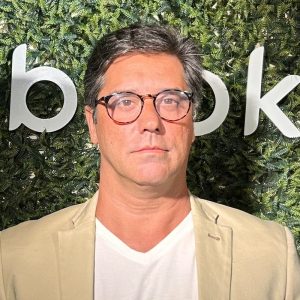 Chris Penna, um homem de pele clara e óculos de aro preto, veste um blazer cinza claro sobre uma camisa branca. Ele está sorrindo levemente e olhando para a câmera. Ao seu fundo, há uma parede verde exuberante coberta por diversas folhagens e o logotipo branco da "bebook" em letras minúsculas e um design circular estilizado. A iluminação natural destaca a cena, sugerindo uma atmosfera profissional e moderna.