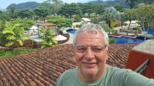 Cláudio Lacerda Oliva, editor da revista Qual Viagem, sorri para uma selfie com cabelos grisalhos e óculos transparentes. Ele está em uma área externa de um hotel ou resort, com um telhado de terracota em primeiro plano e, ao fundo, piscinas, palmeiras e espreguiçadeiras em um ambiente tropical.