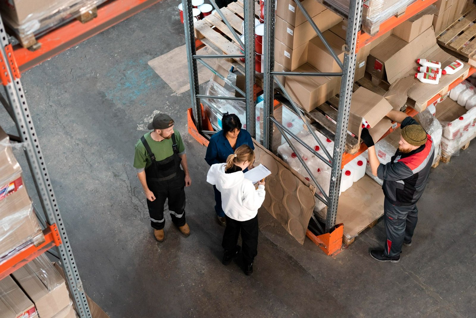 Três funcionários de armazém, dois homens e uma mulher, estão perto de uma prateleira industrial. A mulher está segurando uma prancheta e parece estar orientando a equipe sobre a organização dos produtos, um dos preceitos básicos da implementação da metodologia 5S no estoque. Um dos homens está retirando uma caixa da prateleira, enquanto o outro observa. O armazém parece estar em funcionamento, com o chão de concreto e caixas empilhadas nas prateleiras ao fundo.