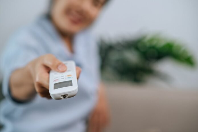Uma mão segura um controle remoto branco apontando para frente, com uma pessoa sorridente e desfocada ao fundo. A imagem ilustra a conveniência da automação, um aspecto-chave da modernização hoteleira.