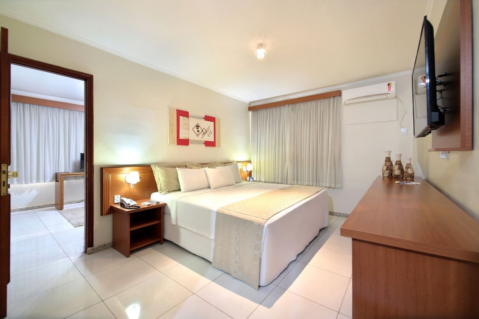 Quarto de hotel com cama king-size, roupa de cama branca e uma peseira (ou "xale de cama") em tom neutro. Há uma mesa de cabeceira de madeira com telefone e luminária ao lado da cama. À frente, um móvel de madeira com uma TV de tela plana pendurada na parede. O chão é de azulejo branco, e uma porta aberta à esquerda leva a um outro cômodo.