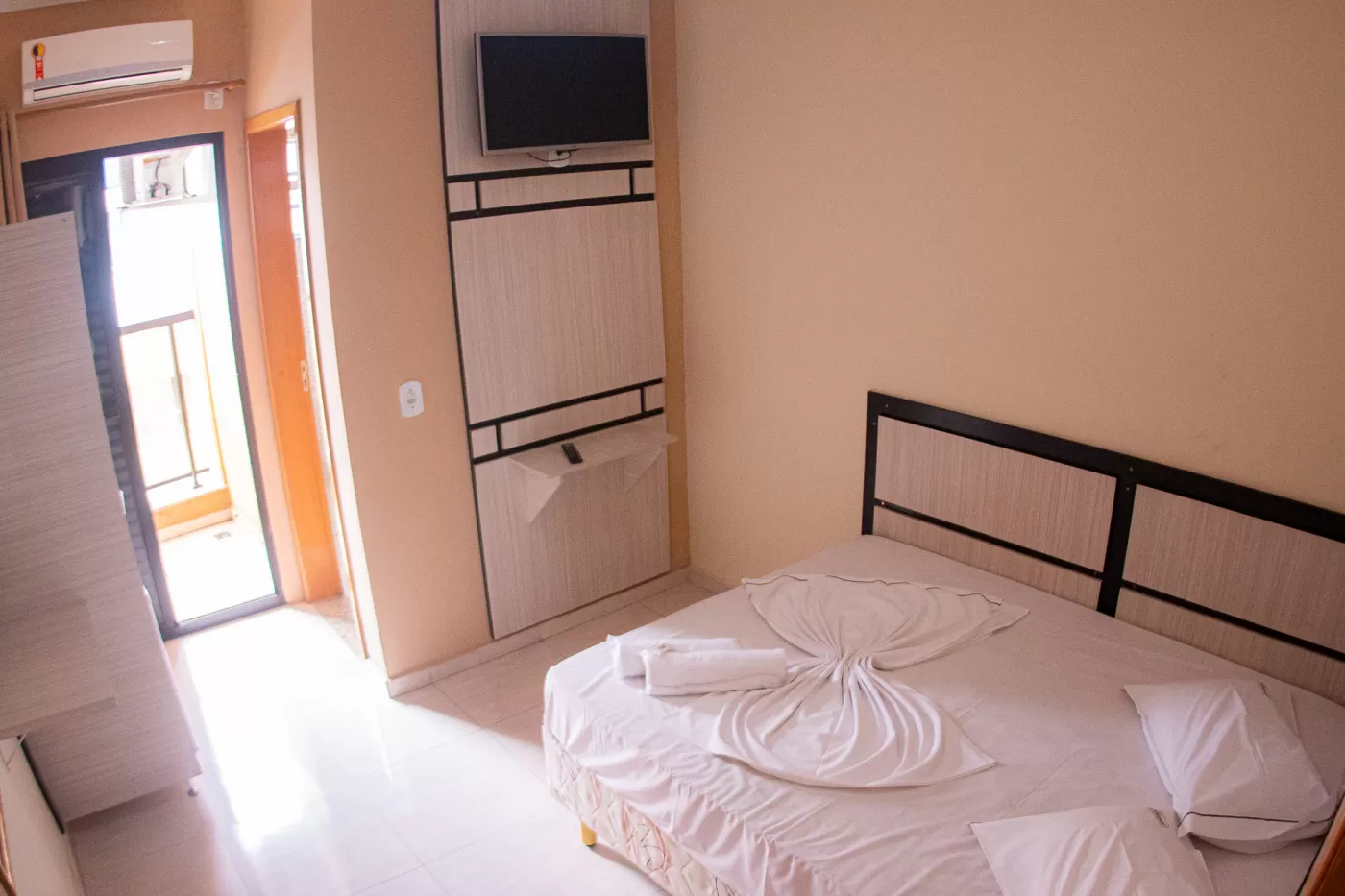 Quarto de hotel simples, com cama de casal e roupa de cama branca e limpa. Os móveis são de cor clara e incluem um painel com TV e um pequeno armário. Ao lado da cama, um controle de remoto está visível. No lado esquerdo, a porta que dá acesso a uma pequena varanda está aberta.