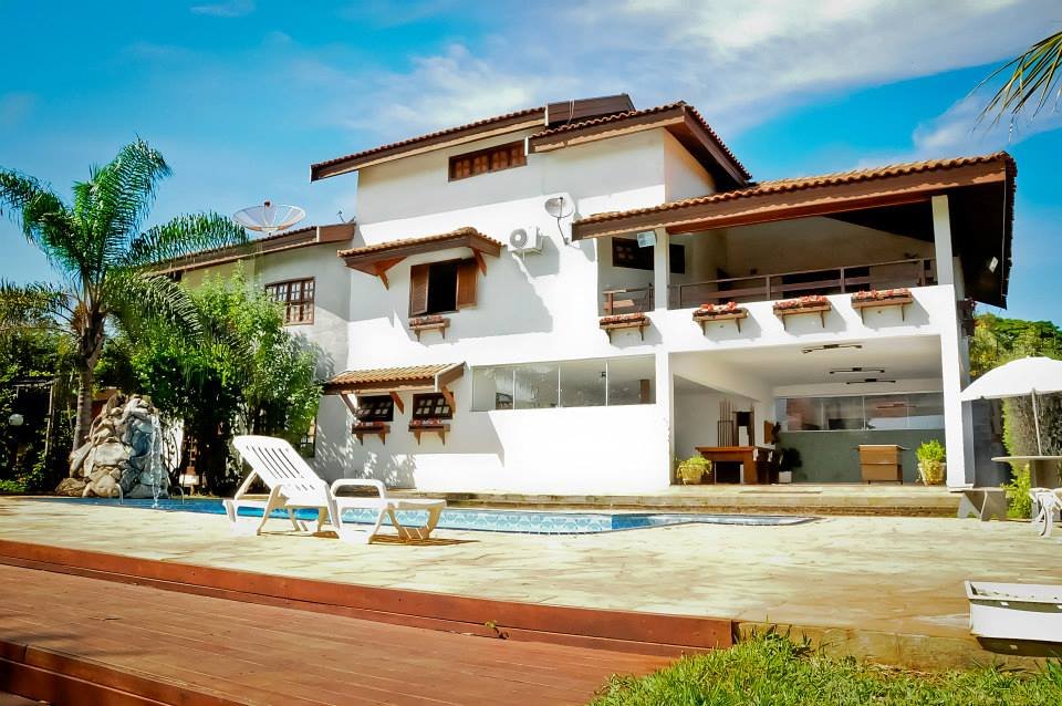 Uma casa grande e branca de dois andares com telhado de telhas e detalhes em madeira. Em frente, há uma piscina retangular e um deck de madeira. Uma espreguiçadeira branca está perto da piscina. À esquerda, uma palmeira alta e folhagens verdes. O céu é azul com algumas nuvens.