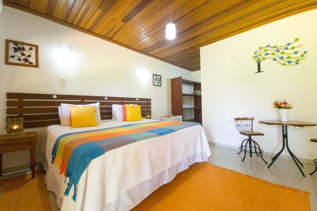 Um quarto de hotel aconchegante com uma cama king-size coberta por um edredom branco, travesseiros amarelos brilhantes e uma manta colorida em tons de azul, laranja e verde. À esquerda da cama, há um criado-mudo de madeira com um abajur rústico. O teto é de madeira escura, e as paredes são brancas, com uma decoração de parede em forma de árvore com folhas coloridas sopradas pelo vento. No canto direito, há uma pequena mesa redonda de madeira com duas cadeiras e um vaso de flores. Um tapete laranja vibrante cobre parte do chão, que é de azulejo claro.