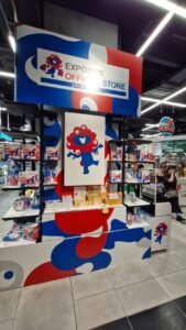Vista interna da loja oficial de produtos da Expo 2025 Osaka. Prateleiras cheias de produtos temáticos com o mascote do evento, Myaku-Myaku, preenchem o espaço. Um grande letreiro no topo anuncia "EXPO 2025 Official Store".