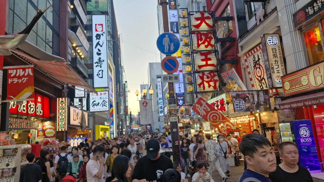Uma rua de Osaka lotada de pedestres. Os prédios ao longo da rua estão cobertos por uma densa e vibrante quantidade de letreiros luminosos e placas com caracteres japoneses. A imagem captura a energia e o movimento de uma área comercial popular da cidade ao anoitecer.