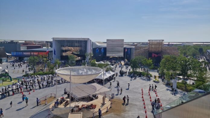 Vista aérea parcial da área externa de um grande complexo de eventos. O espaço é composto por prédios de design moderno, um amplo pátio com pessoas circulando e uma estrutura branca em formato de bacia. A paisagem inclui áreas verdes com árvores e gramados. A imagem retrata a movimentada infraestrutura da Expo 2025 Osaka sob um céu azul.