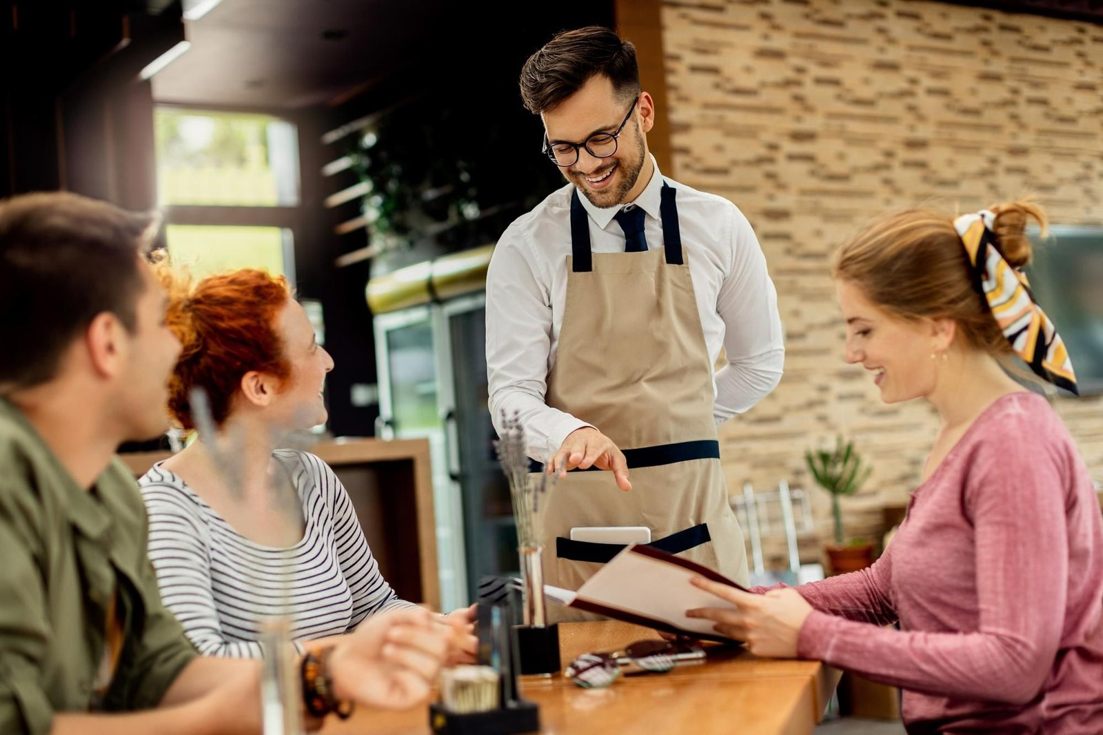 Um garçom sorridente, de óculos, com avental bege e camisa branca, atendendo a uma mesa com três clientes, um homem e duas mulheres. Ele aponta para o cardápio que uma das mulheres segura, enquanto os outros clientes olham para ele com expressão satisfatória. 