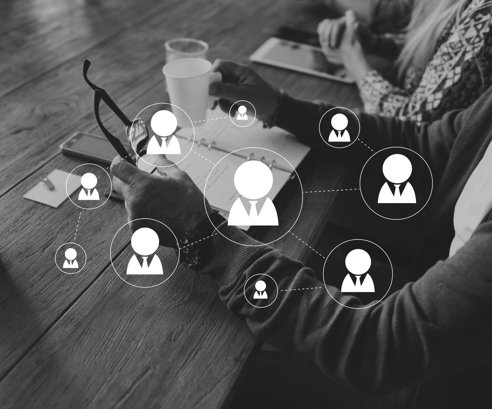 Uma foto em preto e branco de uma mesa de reunião com pelo menos duas pessoas. O foco está nas mãos de um profissional, que segura os óculos enquanto aponta para um caderno aberto. Sobre a imagem, há uma sobreposição digital com ícones de pessoas conectados por linhas pontilhadas, representando o conceito de coletividade. 